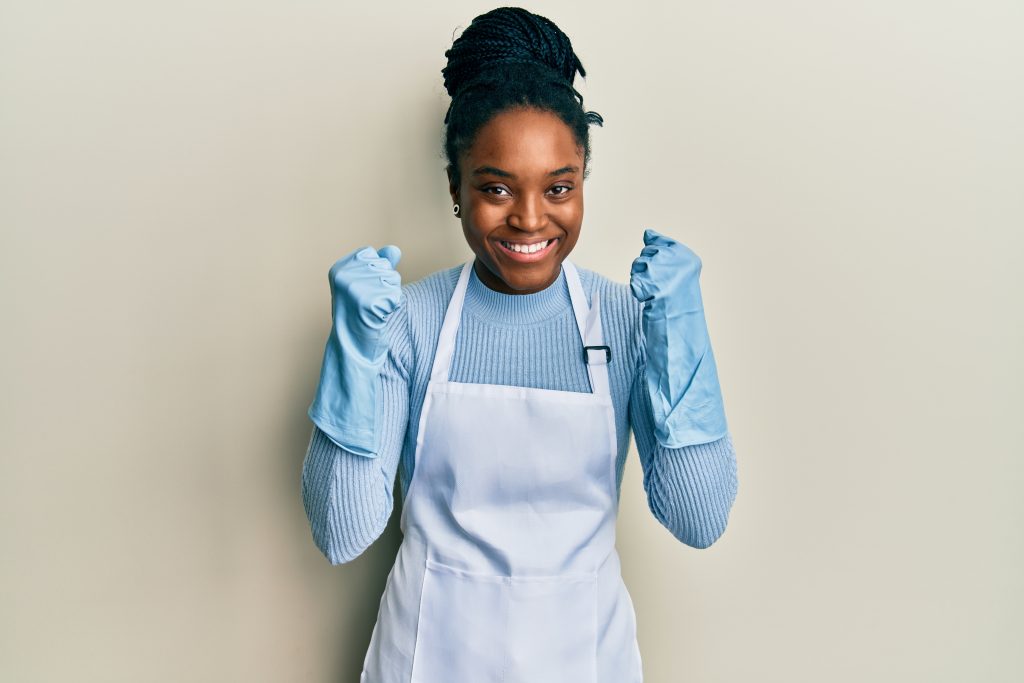 african american woman with braided hair wearing cleaner apron and gloves celebrating surprised and amazed for success with arms raised and open eyes. winner concept.